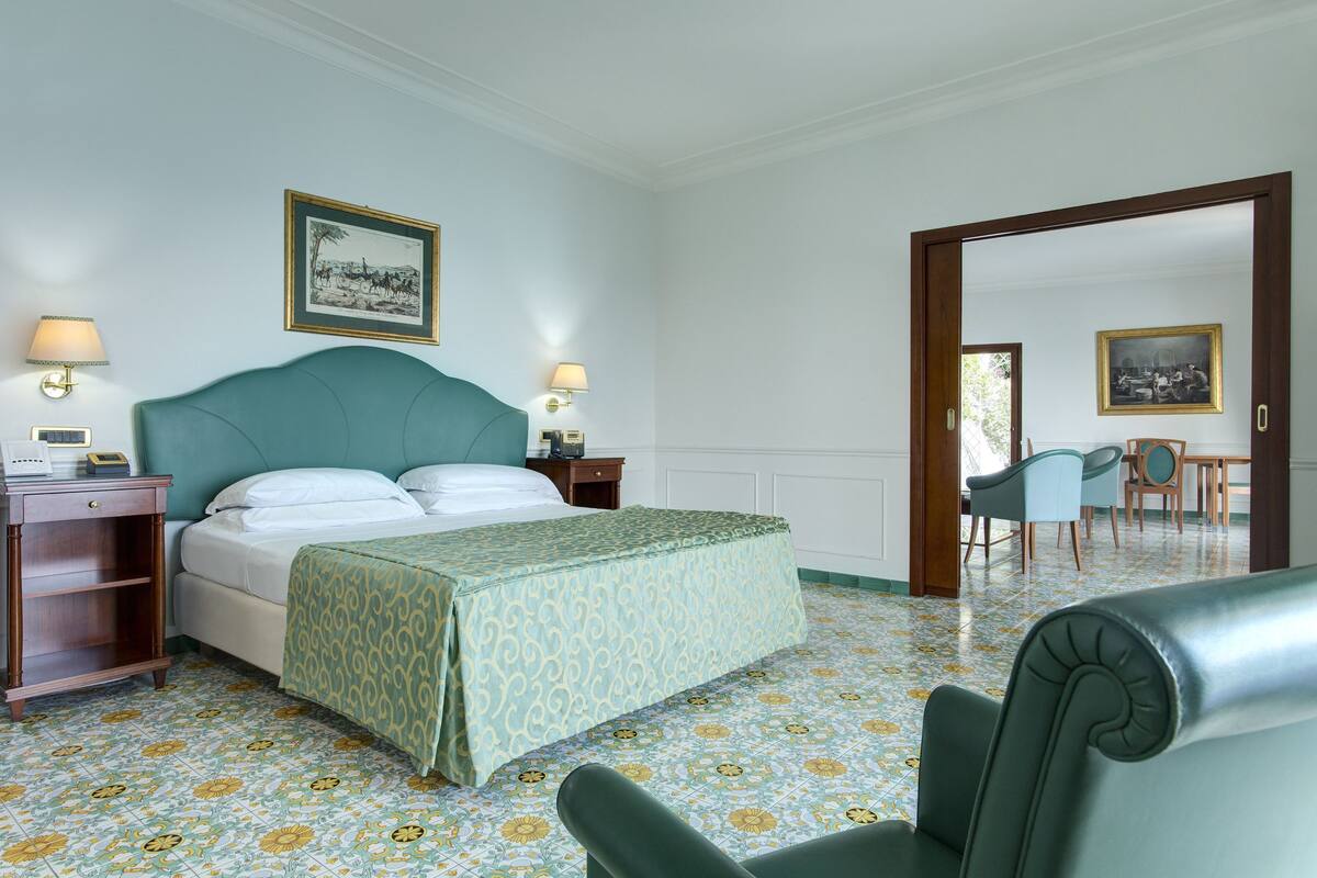 Habitació d'hotel amb un llit, dues taules de nit amb làmpades, una cadira i una zona de menjador amb cadires i taula.