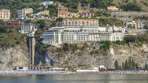 Un hotel frente al mar con varios pisos, una playa en primer plano y una ladera rocosa al fondo.