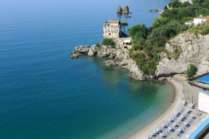 Un hôtel en bord de mer, situé sur une falaise, avec une plage équipée de parasols et un bâtiment historique.