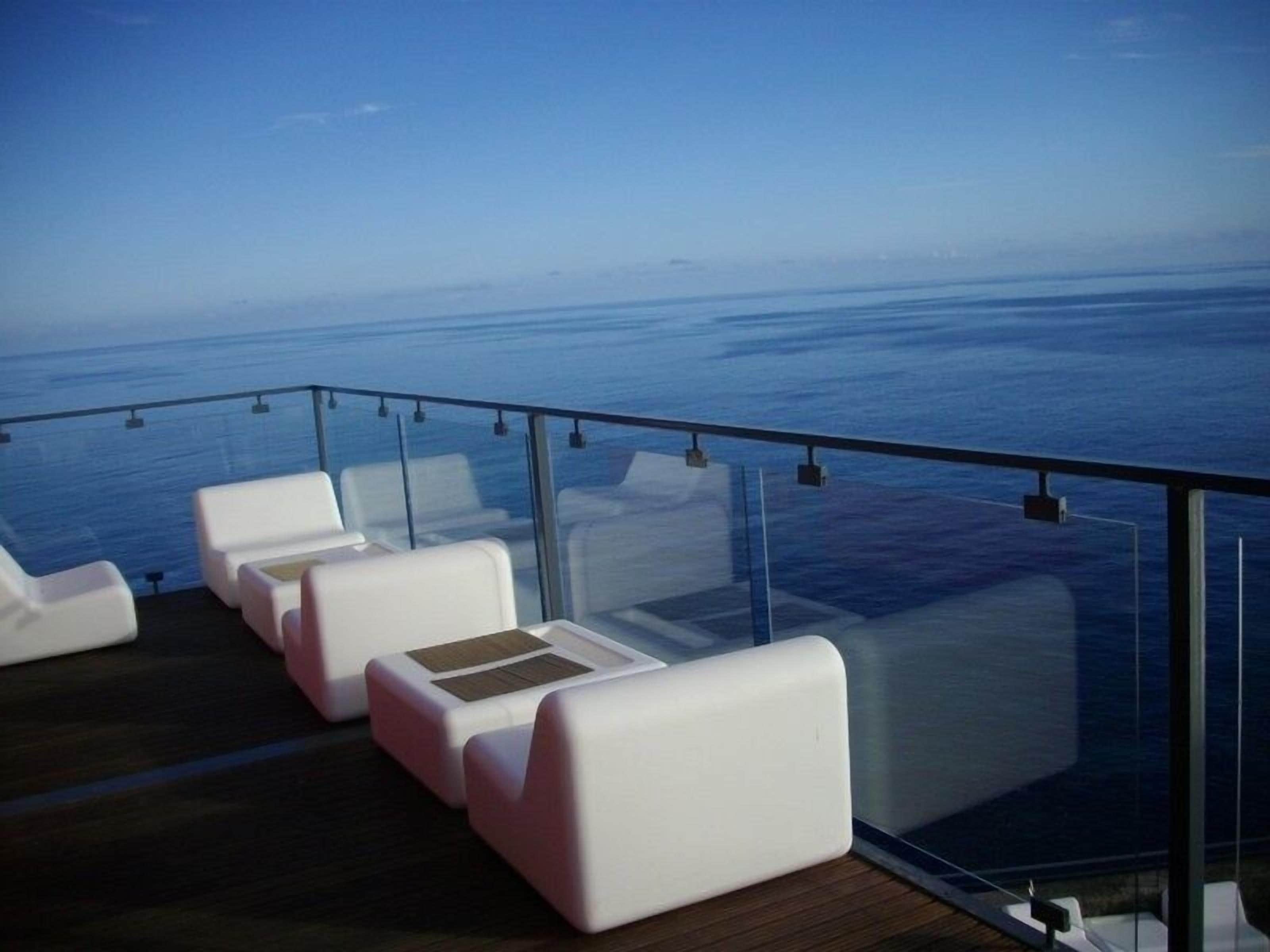 A deck with white cushioned seating and a glass railing overlooking the sea.
