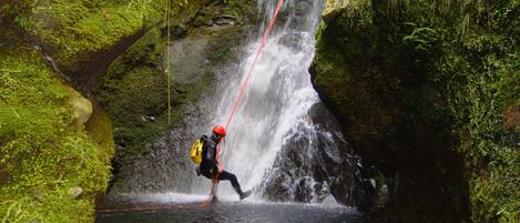 A person rappelling down a waterfall in a natural setting with moss-covered rocks and a clear water pool at the bottom.