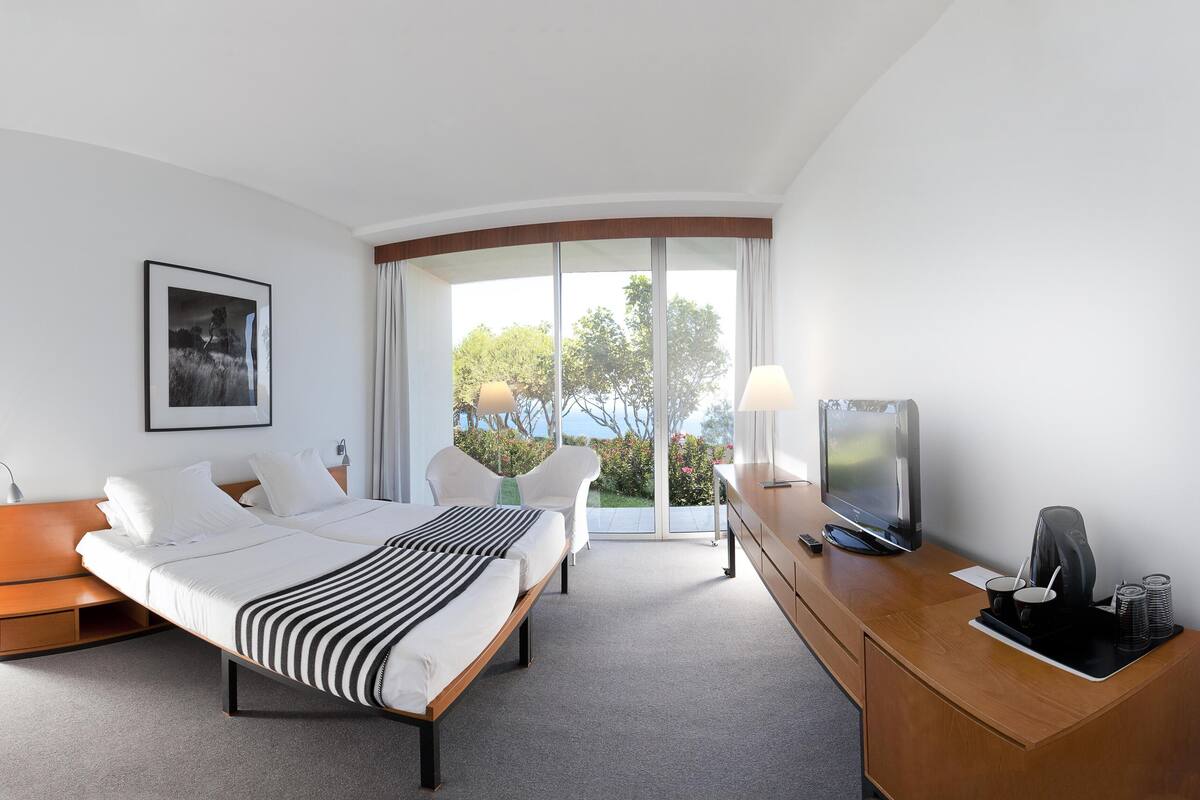 Une chambre d’hôtel moderne équipée d’un lit, d’un bureau avec une télévision et d’une grande fenêtre donnant sur les arbres et le ciel.
