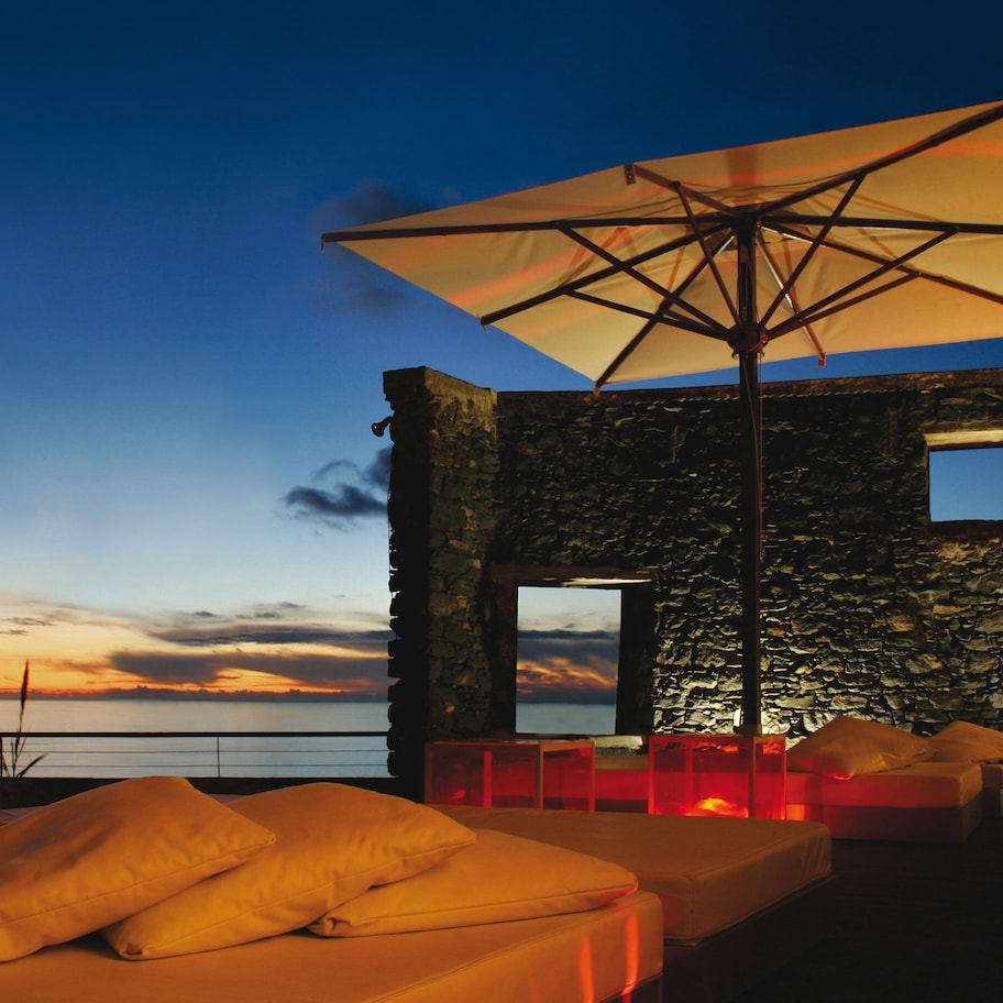 A stone building with a bed and an umbrella, overlooking a sunset.