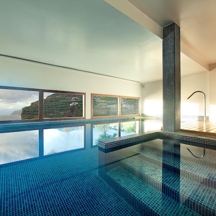 A modern indoor swimming pool with clear glass walls, a view of the sea, and a tiled floor.