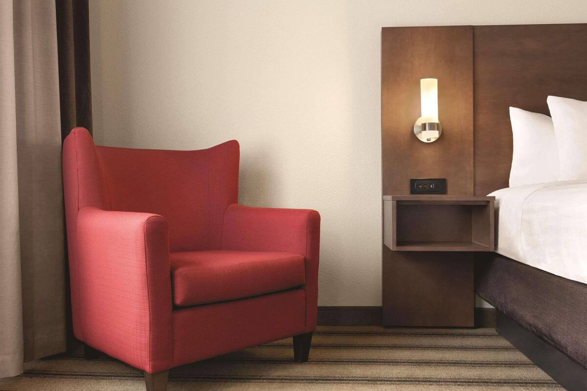 A hotel room with a red armchair, a bedside table with a lamp, and a bed with white linens.