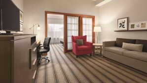 A hotel room with a TV, a desk, a red armchair, a sofa, and a window with curtains. - Country Inn & Suites by Radisson, Billings, MT (Billings)