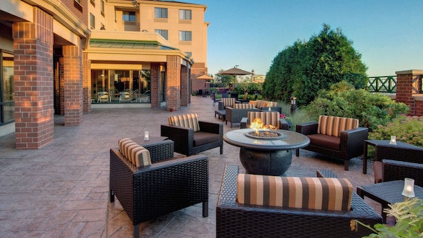 An outdoor patio area with wicker furniture, a central fire pit, and a building with a brick facade in the background. - Courtyard by Marriott Madison East (Madison)