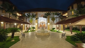 A courtyard with a central fountain, surrounded by palm trees and benches, leading to a building with multiple balconies and a thatched roof. - Occidental Caribe All Inclusive (Punta Cana)