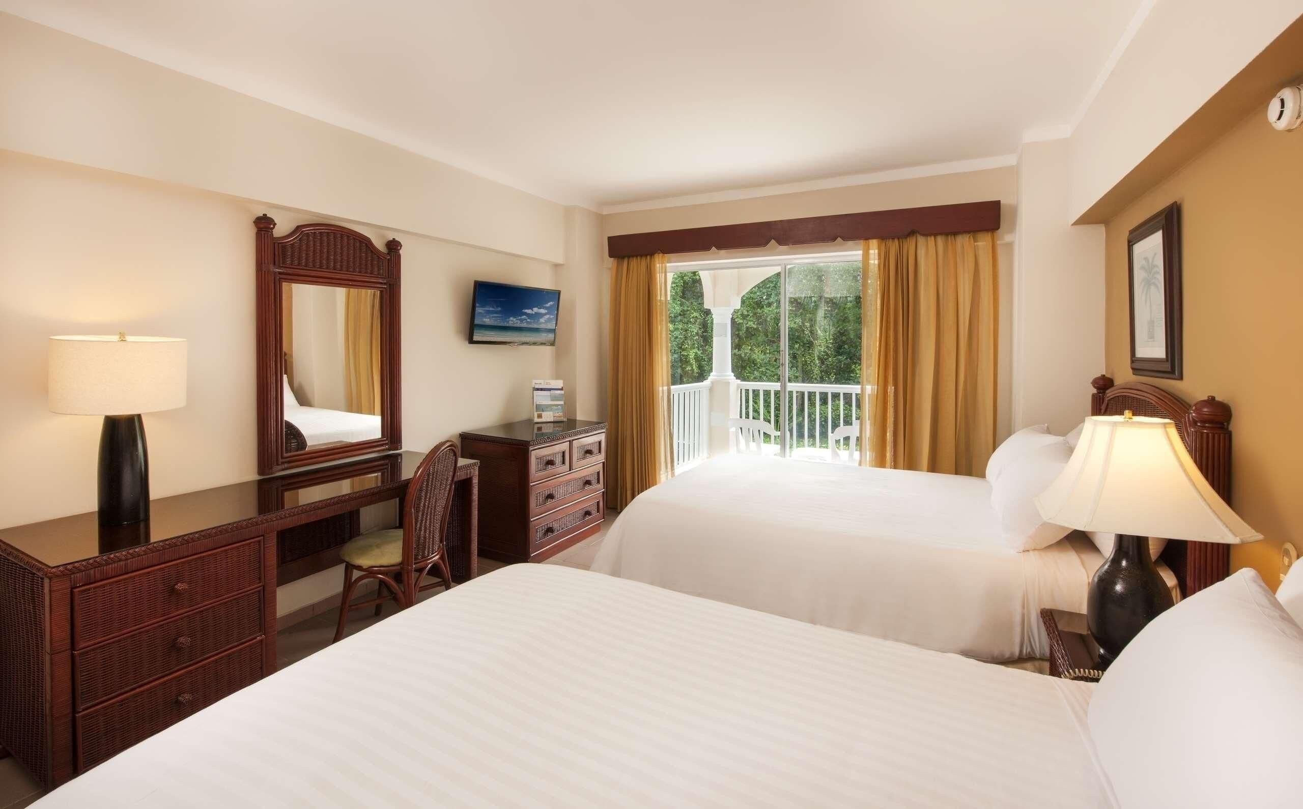A hotel room with two beds, a desk with a chair, a dresser, a TV, and a balcony with curtains.