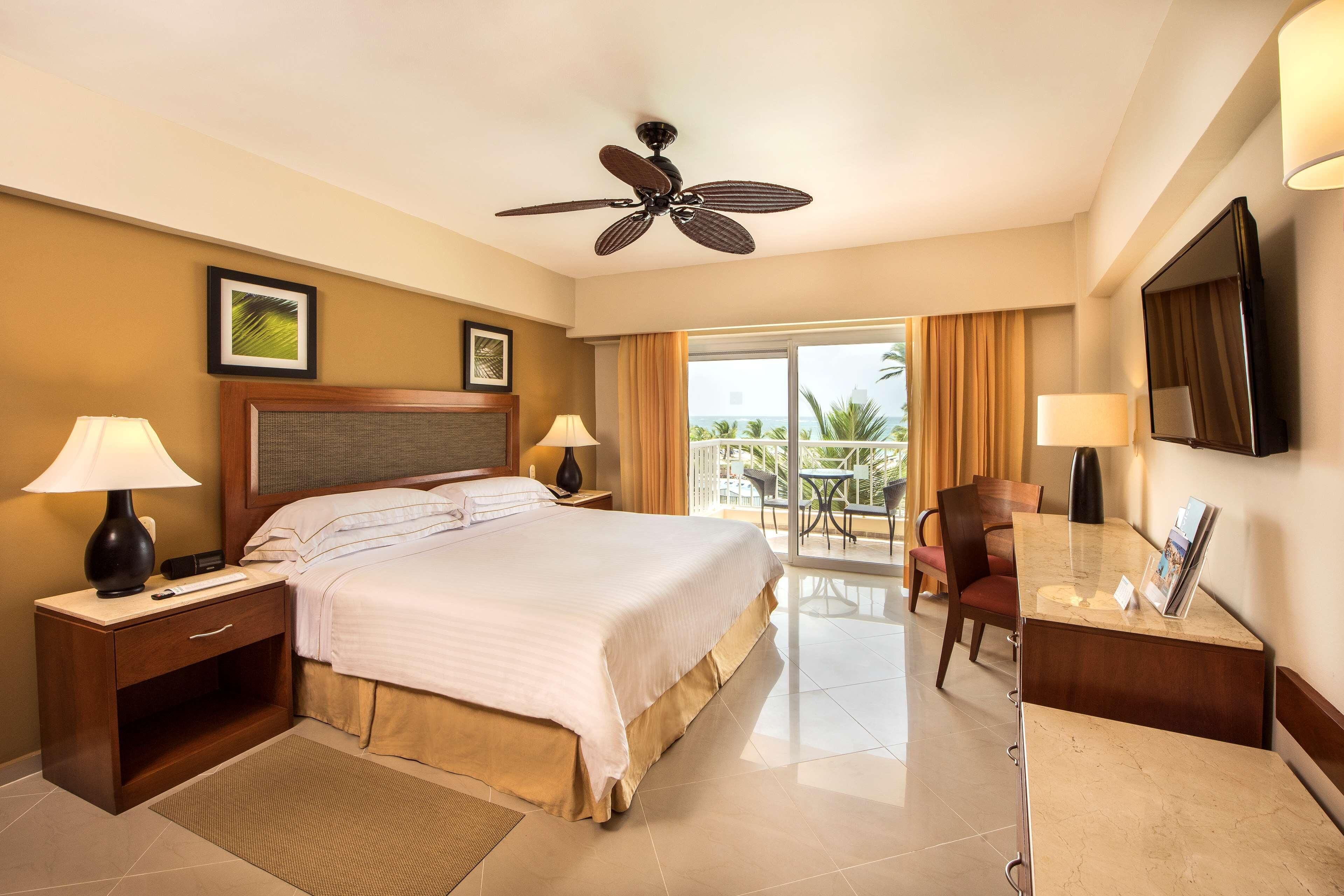 A hotel room with a large bed, two bedside tables with lamps, a ceiling fan, a desk with a chair, and a balcony with a view.