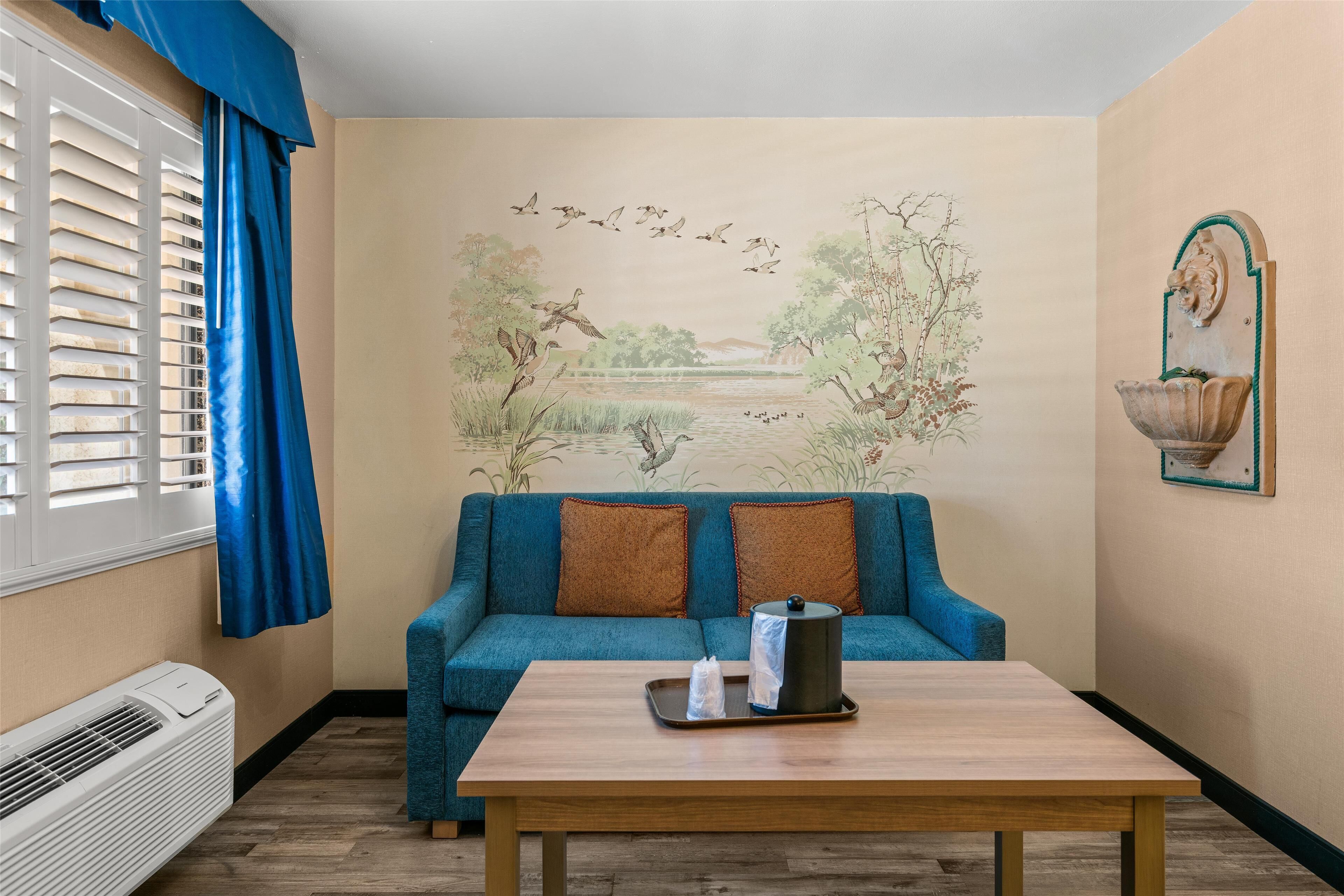 A room with a blue sofa, a wooden table, a wall mural, and a window with blinds.