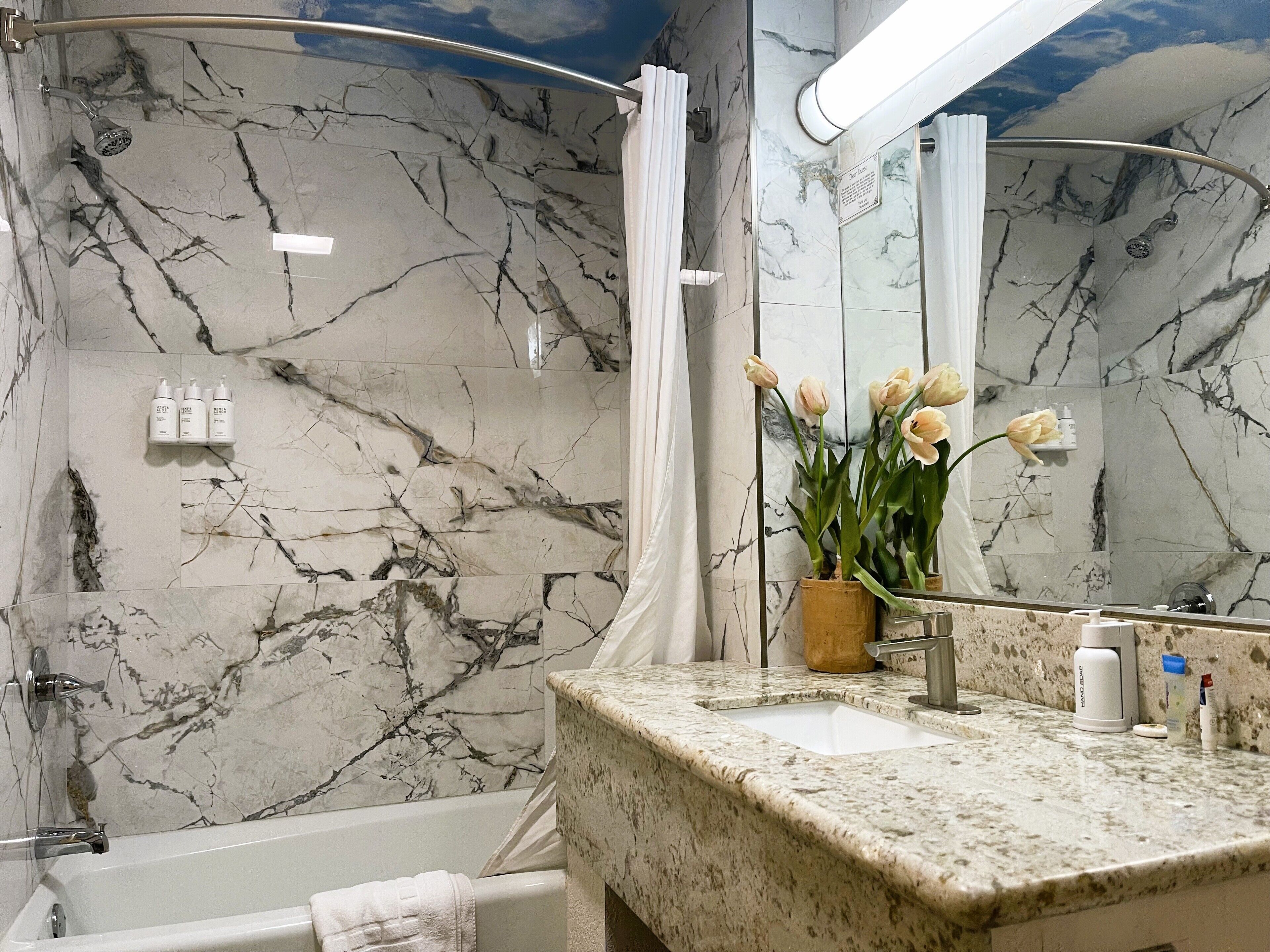 A bathroom with a marble wall, a bathtub, a shower with a curved glass door, a sink with toiletries, and a mirror reflecting the bathroom.