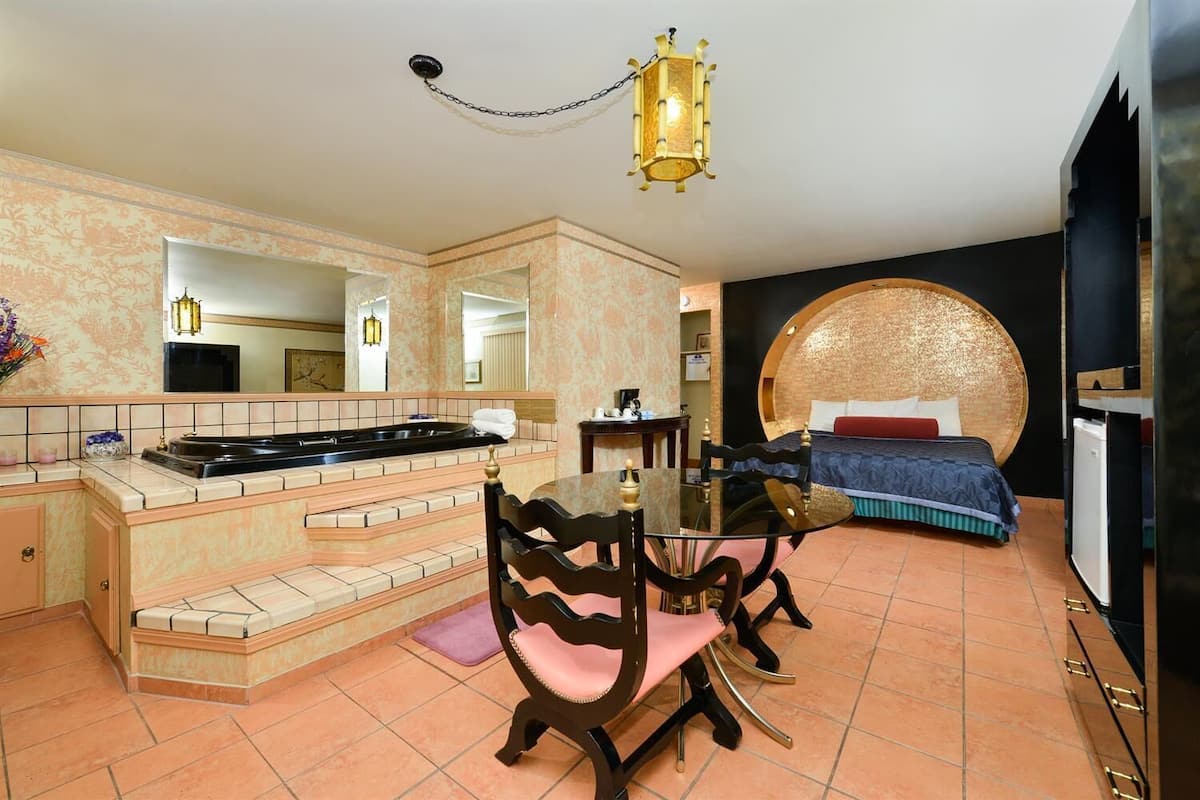 Habitación de hotel con cocina, mesa de comedor con sillas, cama con cabecero original y un espejo grande.