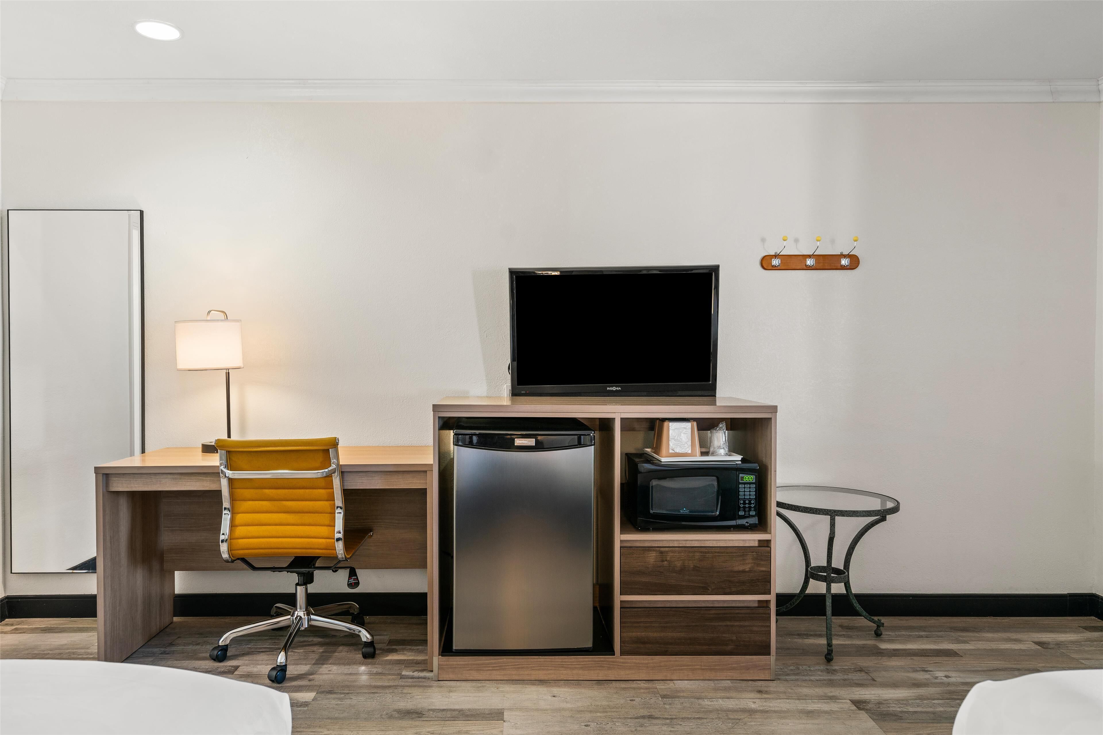 A hotel room with a wooden desk, a yellow office chair, a refrigerator, a microwave, a television, a small round table, and a wall-mounted light fixture.