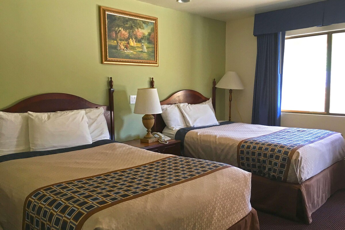 A hotel room with two beds, a painting on the wall, a lamp, and a window with curtains.