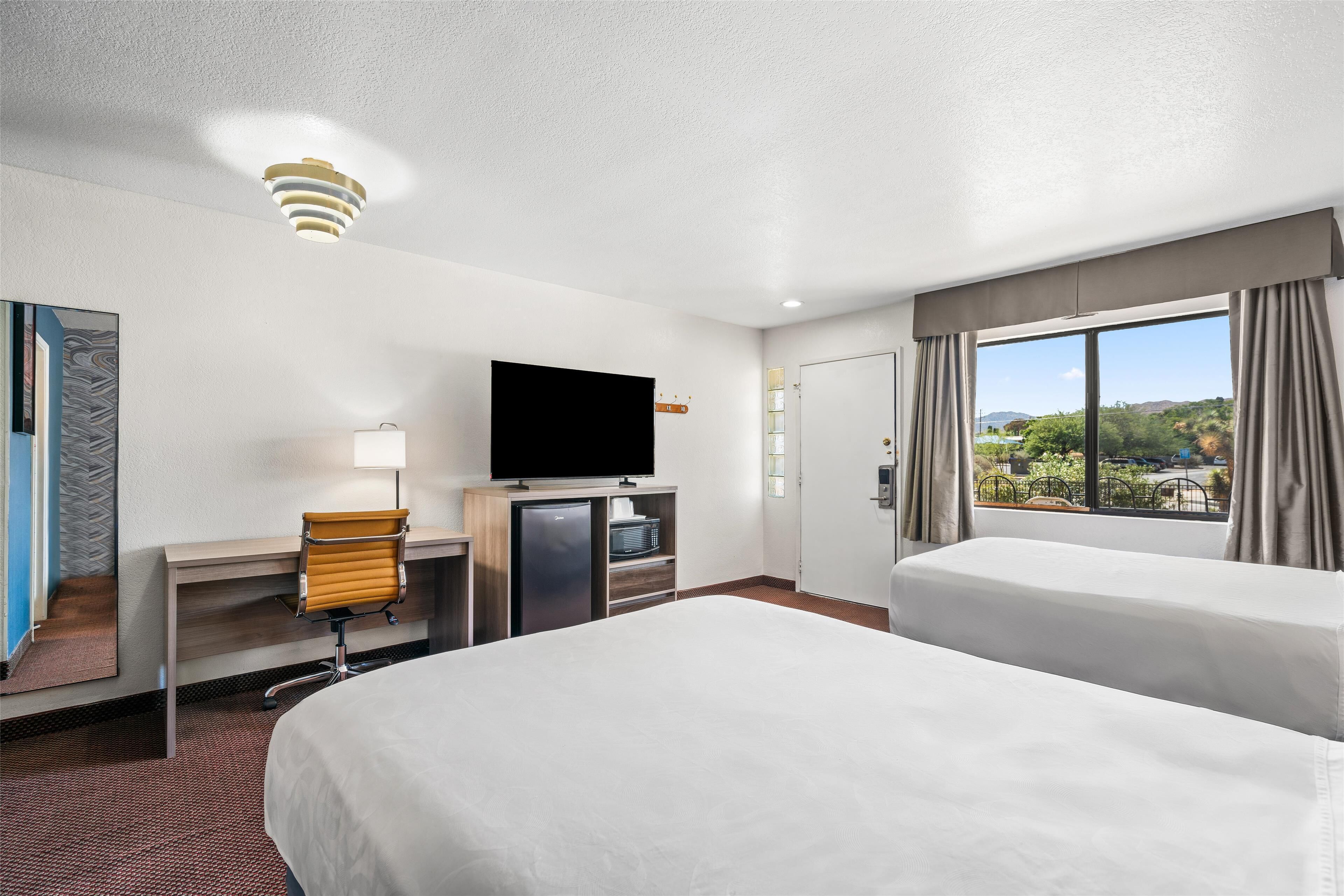 A hotel room with two beds, a desk with a TV, a minibar, and a window with a view of greenery.
