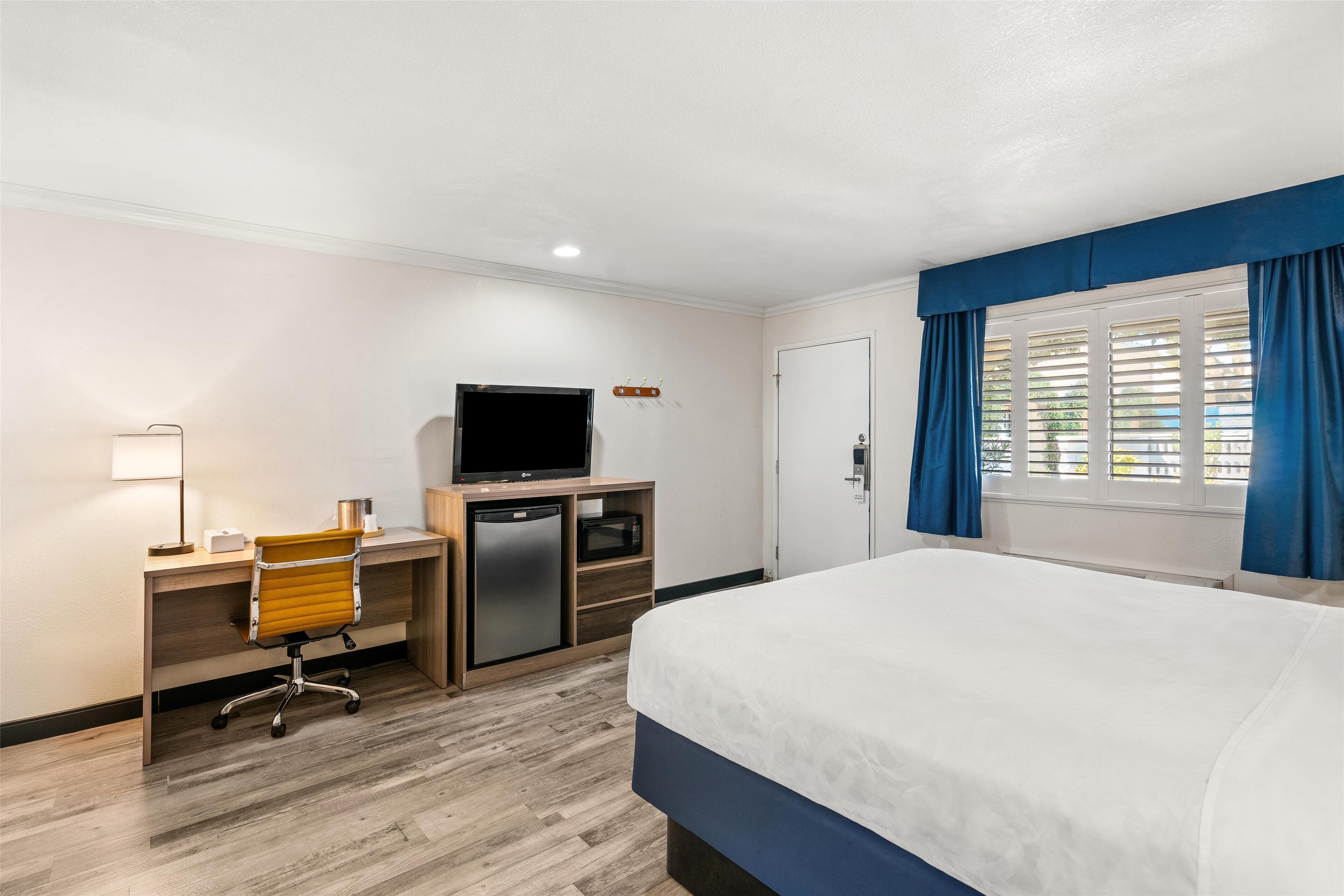 Una habitación de hotel con cama, escritorio, silla, televisor y ventana con cortinas.