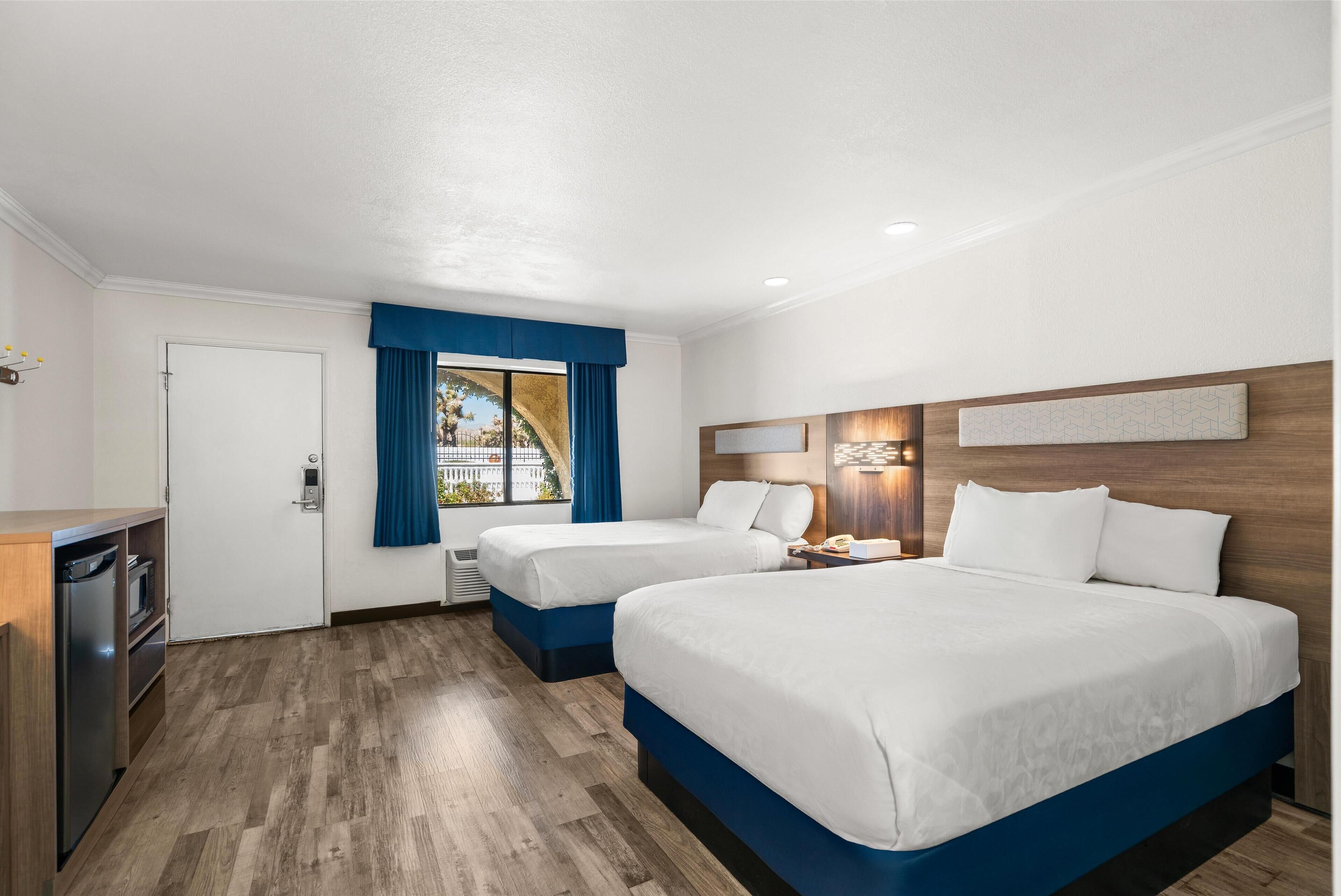A hotel room with two beds, a desk, and a window with blue curtains.