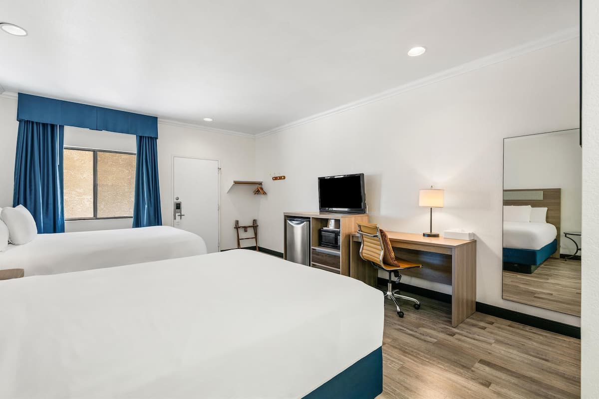 Habitación de hotel con dos camas, un escritorio con silla, televisor, espejo y ventana con cortinas.