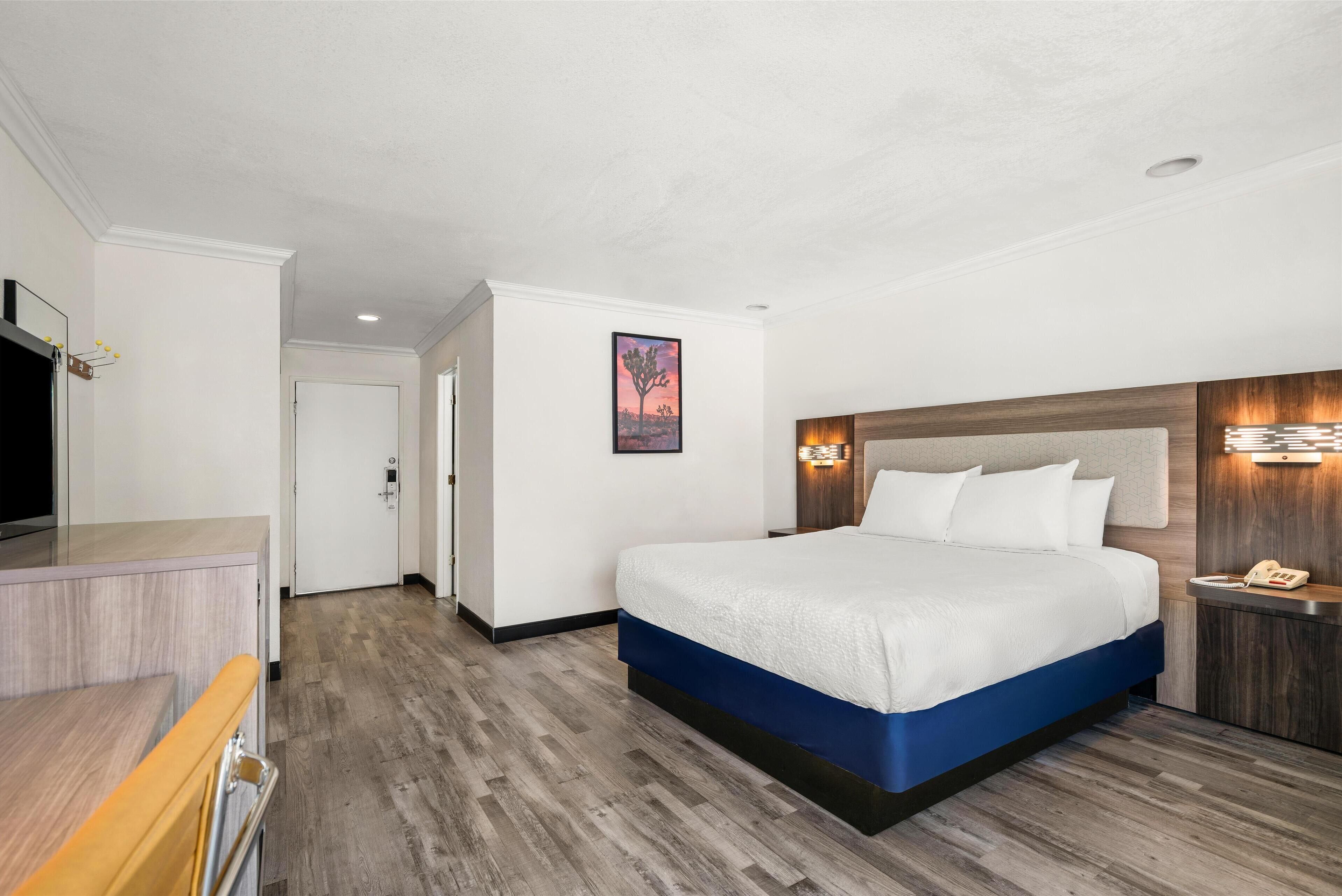 A hotel room with a large bed, a wooden headboard, a bedside table with a lamp, and a flat-screen TV.