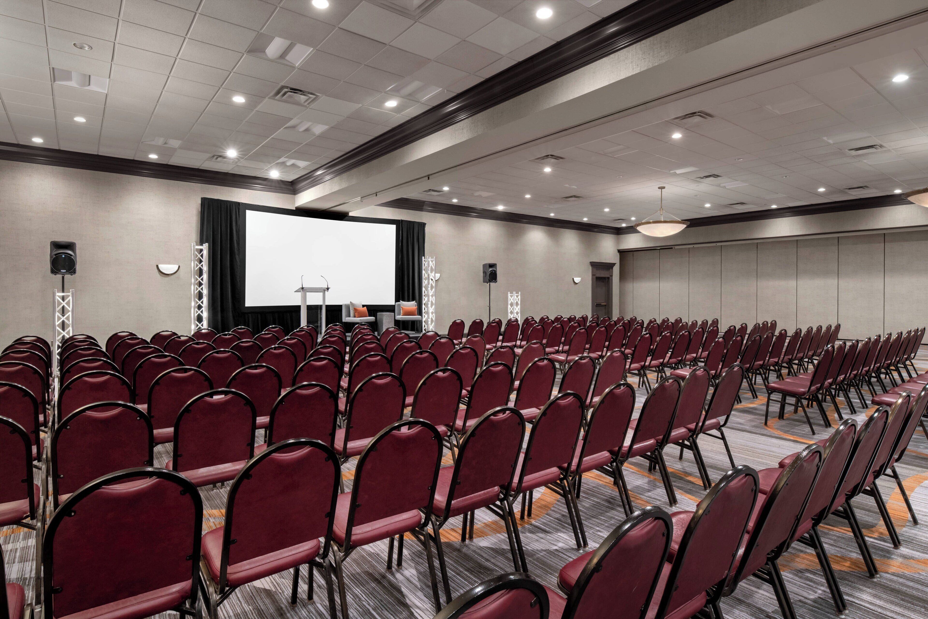 A conference room with rows of red chairs, a large screen, and a podium.
