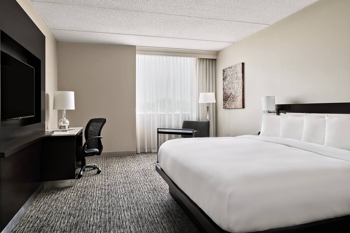 A hotel room with a large bed, a desk with a chair, a flat-screen TV, and a window with curtains.