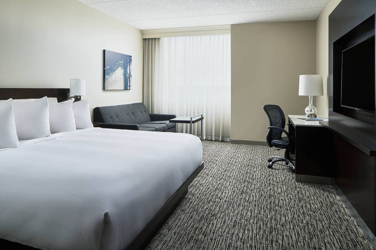 A hotel room with a large bed, a desk with a chair, a flat-screen TV, and a window with curtains.