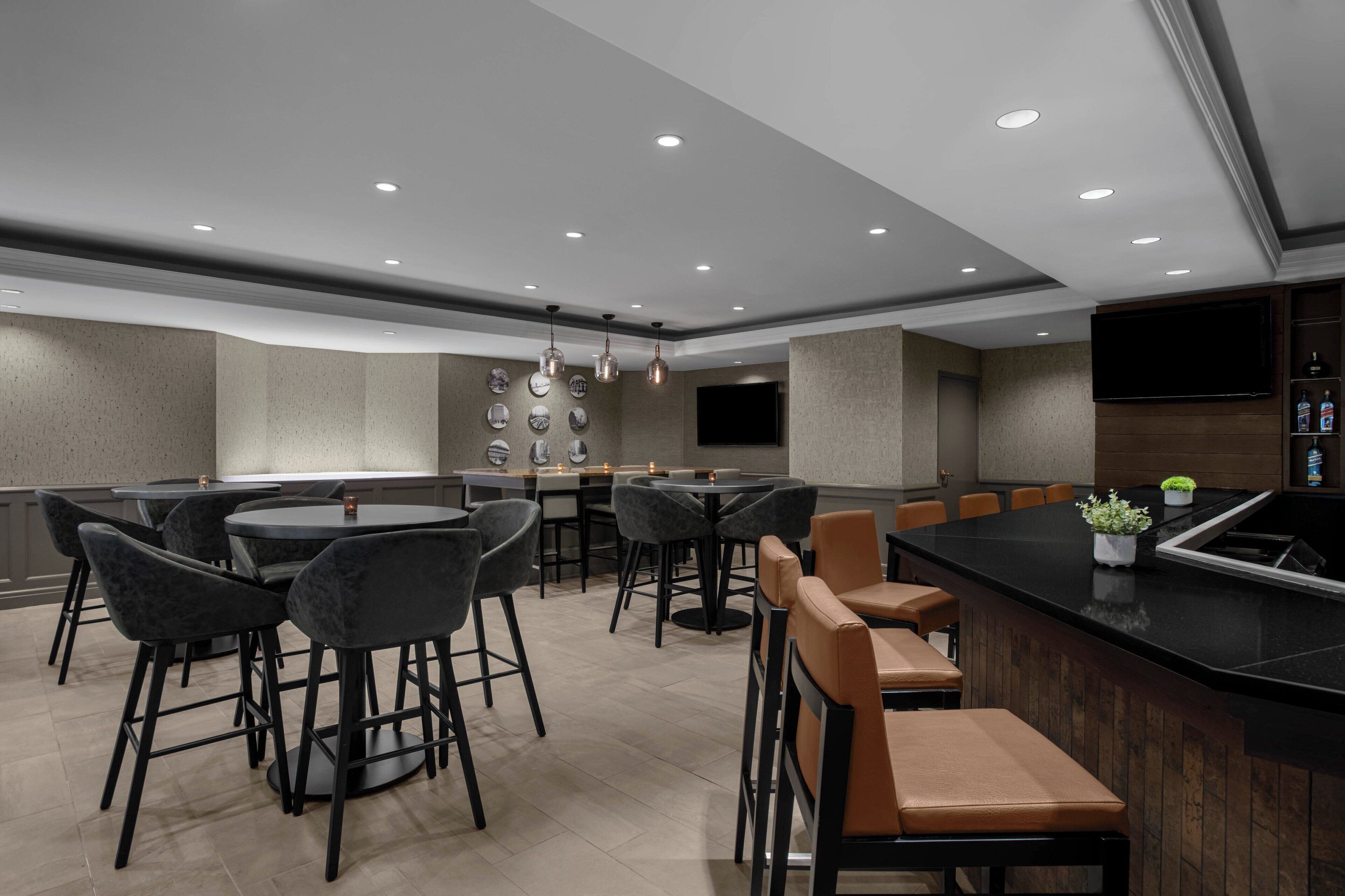Un área de bar moderna con mesas redondas y taburetes de bar, un televisor de pantalla plana en la pared y una variedad de platos decorativos en la pared.