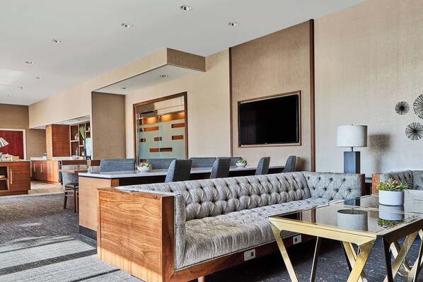 Un hall d’hôtel moderne doté d’un espace salon, d’un comptoir d’accueil en bois et d’un grand téléviseur à écran plat.