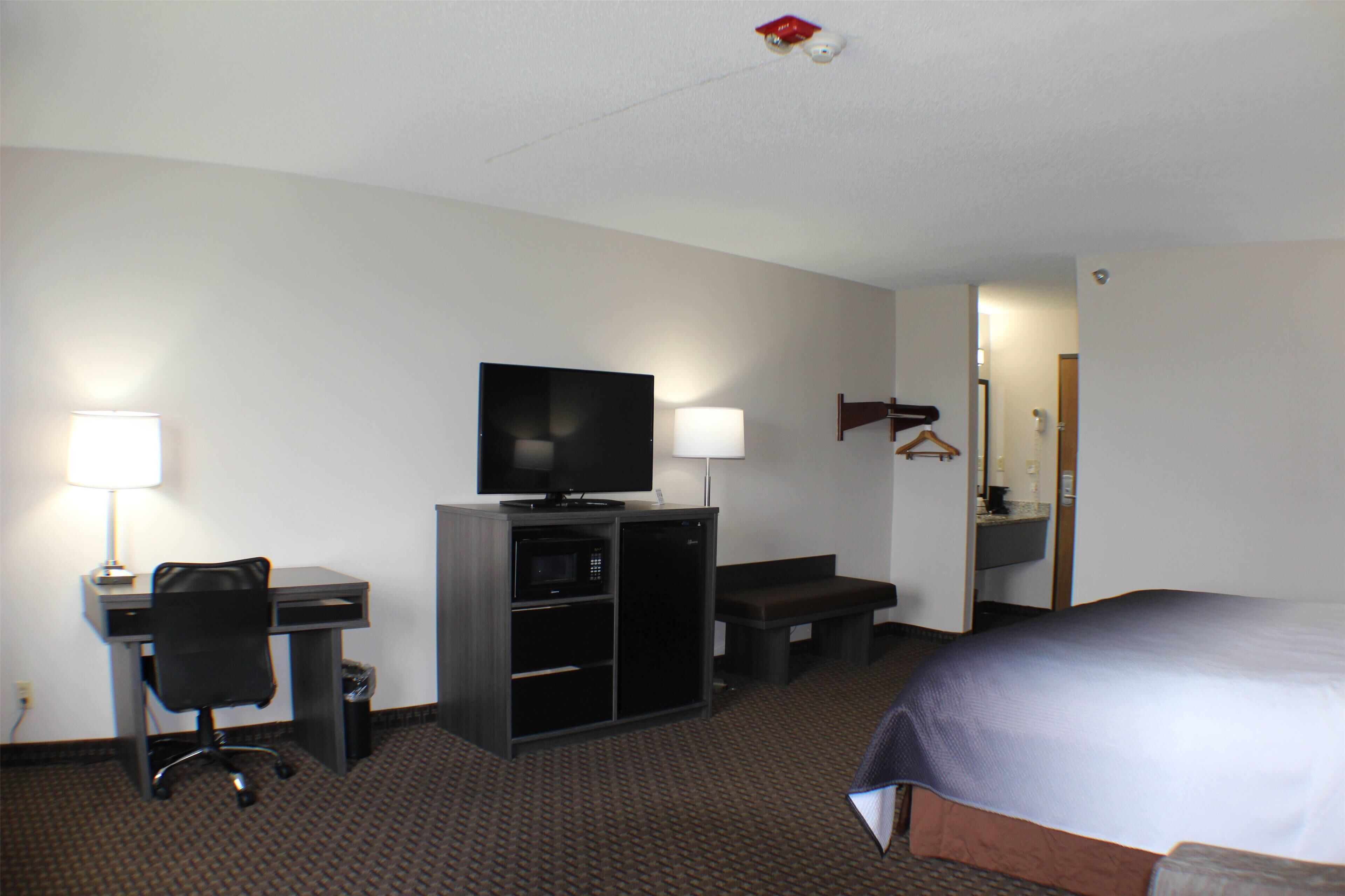 A hotel room with a bed, a desk with a computer, a television, a lamp, a bench, and a bathroom.