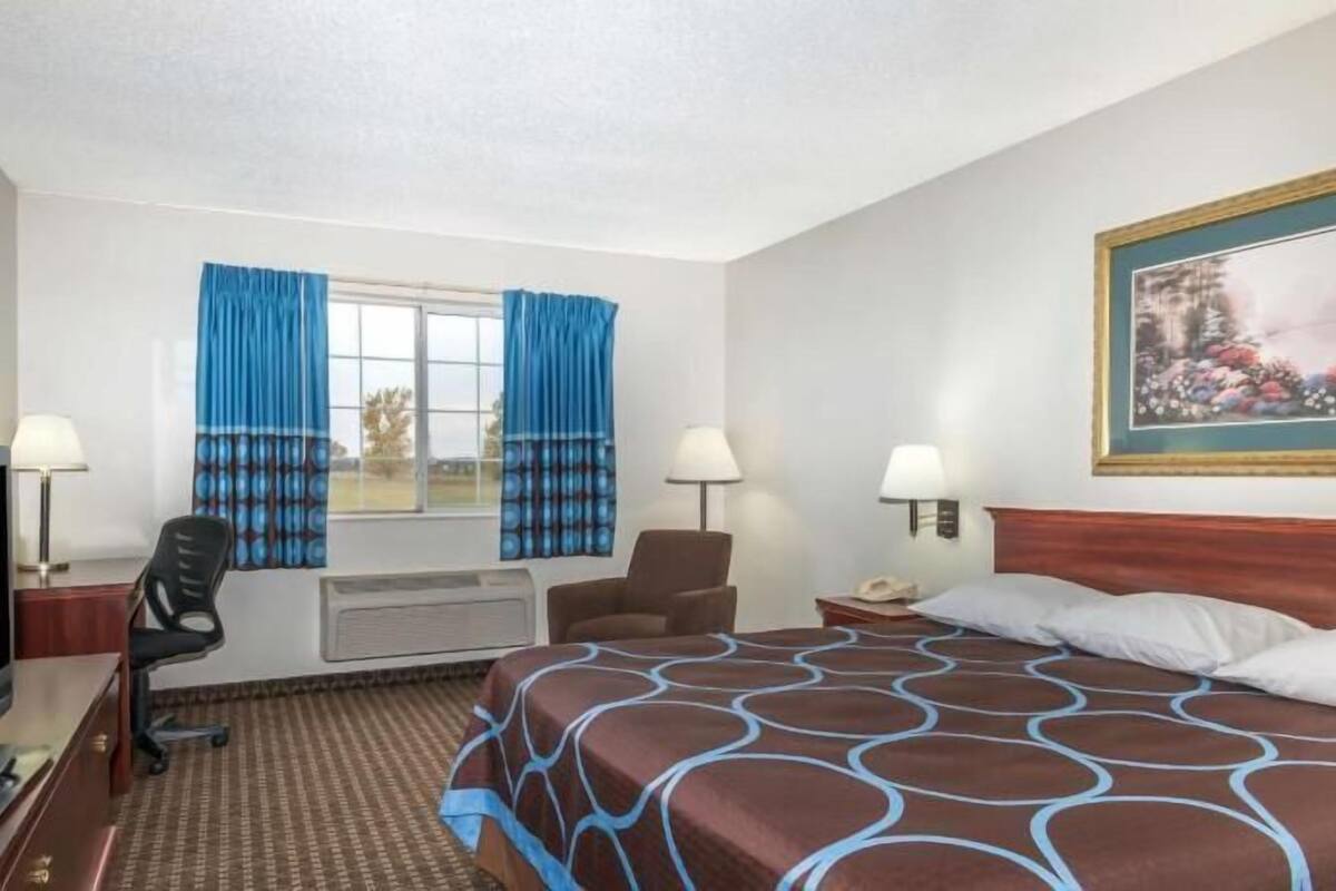 Een hotelkamer met een bed, een bureau met stoel, een televisie, een raam met gordijnen, een schilderij aan de muur en een lamp.