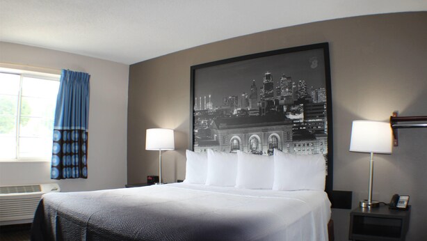 Une chambre d’hôtel avec un grand lit, deux lampes de chevet, une œuvre d’art représentant un paysage urbain accrochée au mur, une fenêtre avec des rideaux bleus et un radiateur.