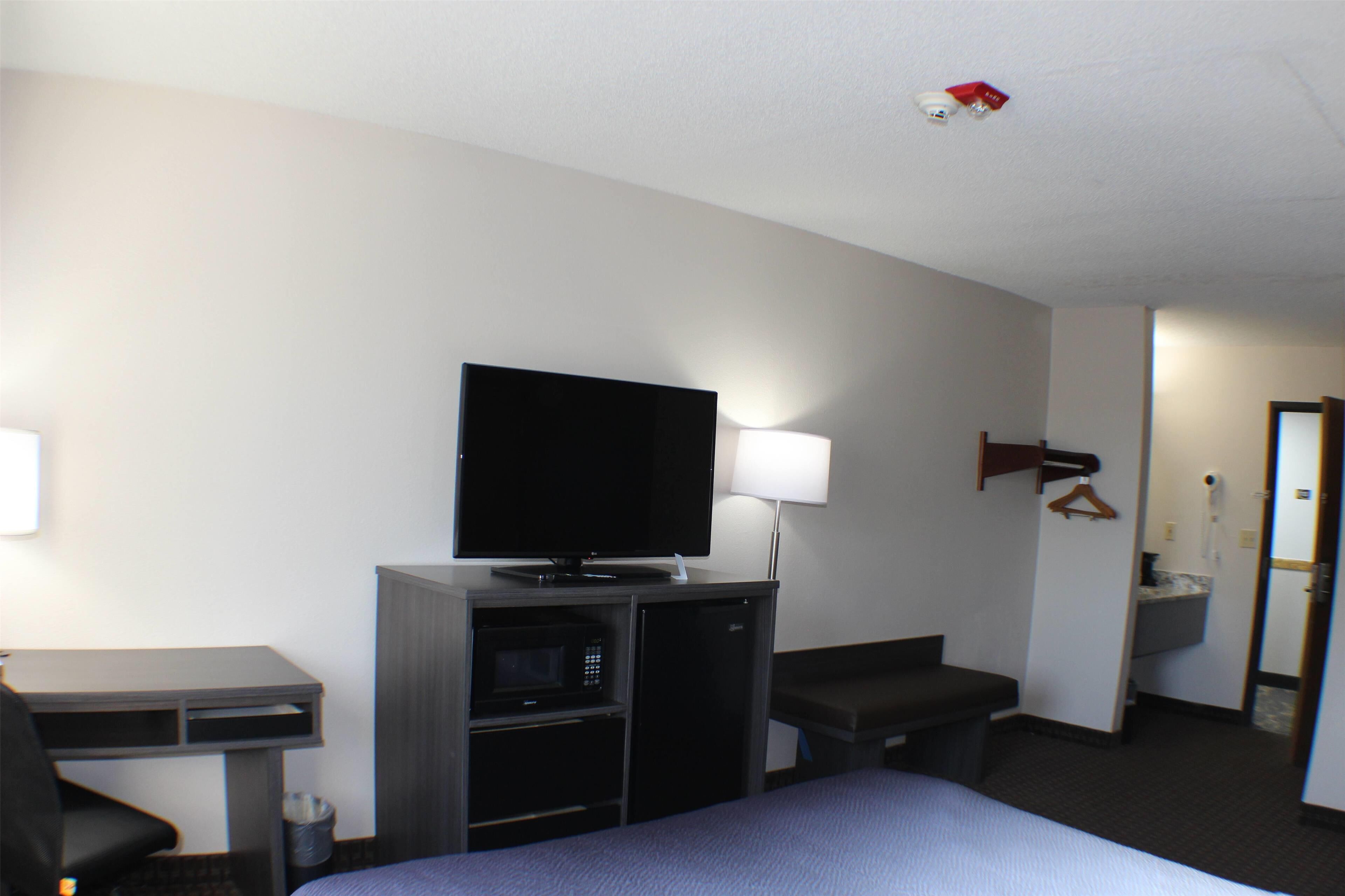 A hotel room with a bed, a desk with a computer, a flat-screen TV, a microwave, a lamp, and a bathroom visible through an open door.