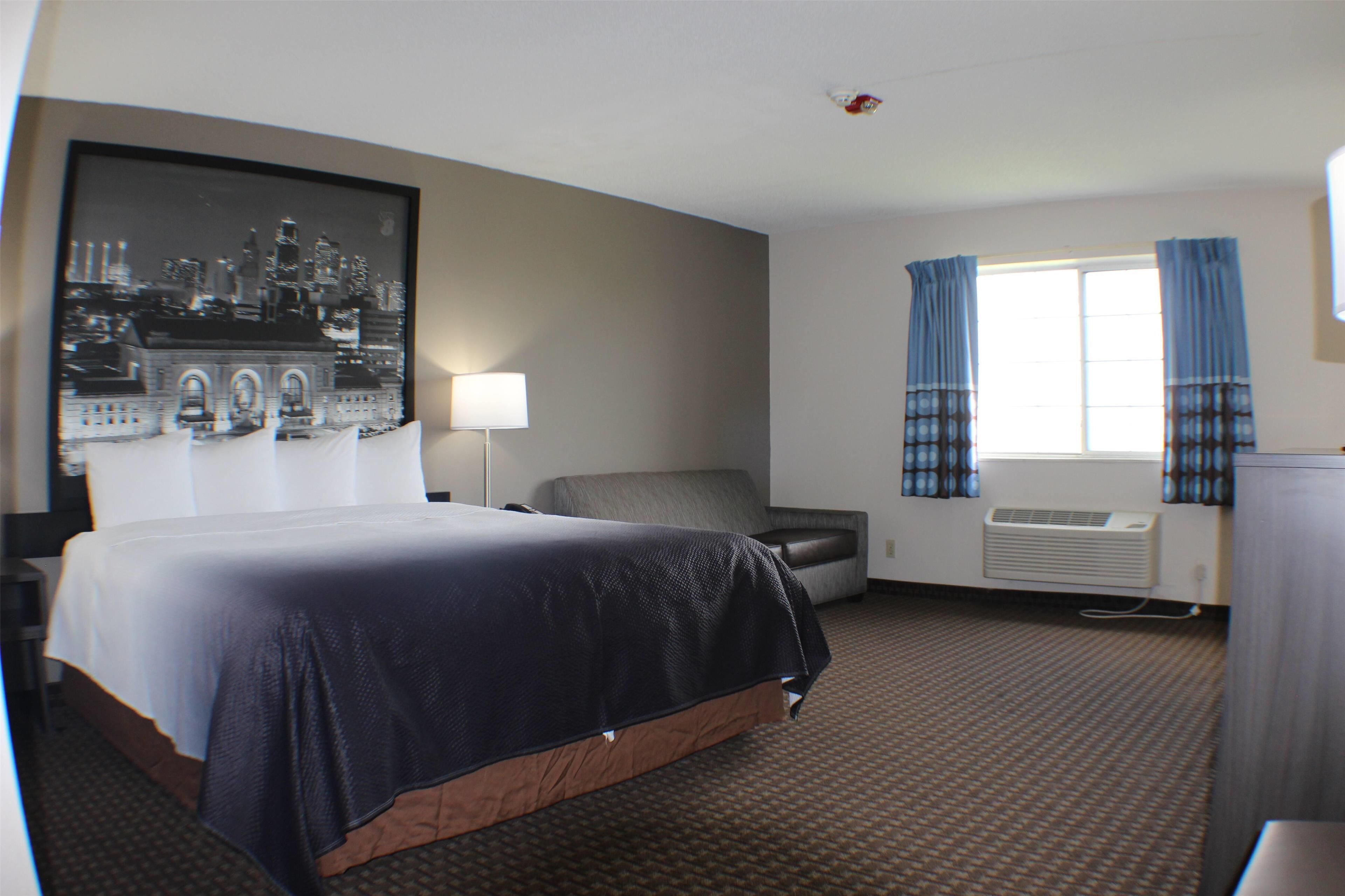 A hotel room with a large bed, a chair, a desk, a lamp, a window with curtains, and a wall-mounted air conditioner.