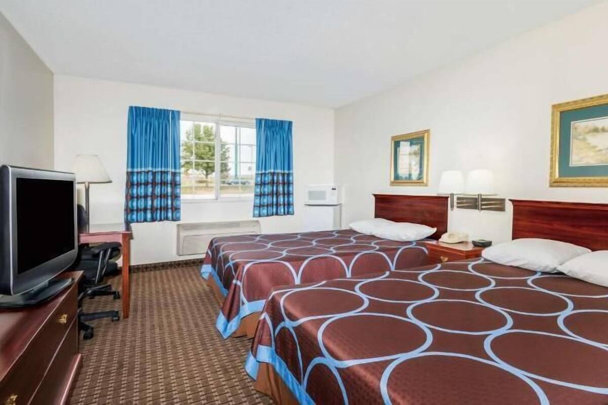 Une chambre d’hôtel équipée d’une télévision, de deux lits avec une literie à motifs, d’un bureau avec une lampe et d’une fenêtre avec des rideaux.