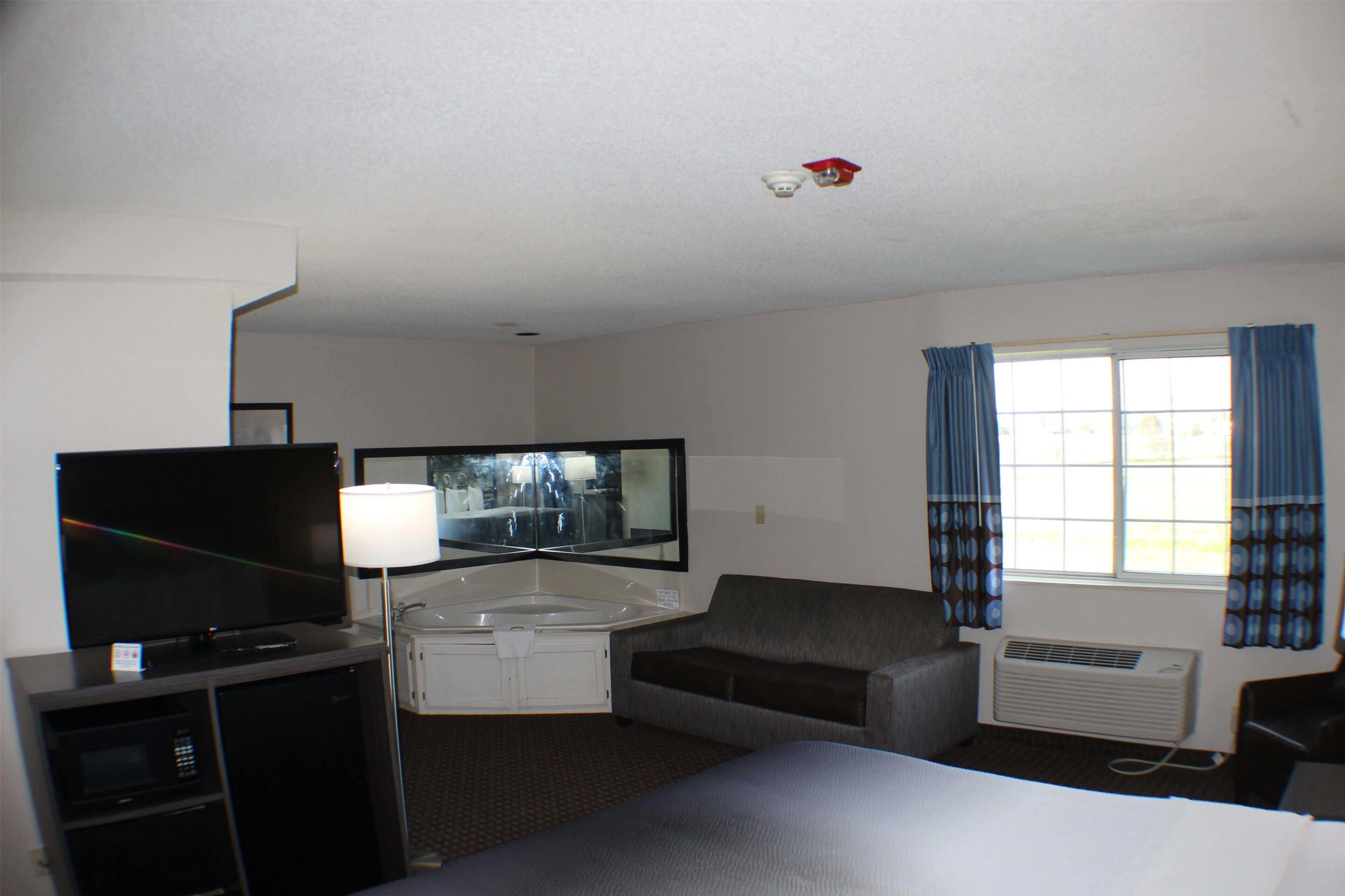 A hotel room with a bed, a desk with a computer, a television, a sofa, a window with curtains, and an air conditioning unit.
