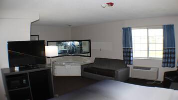 A hotel room with a bed, a desk with a computer, a television, a sofa, a window with curtains, and an air conditioning unit.