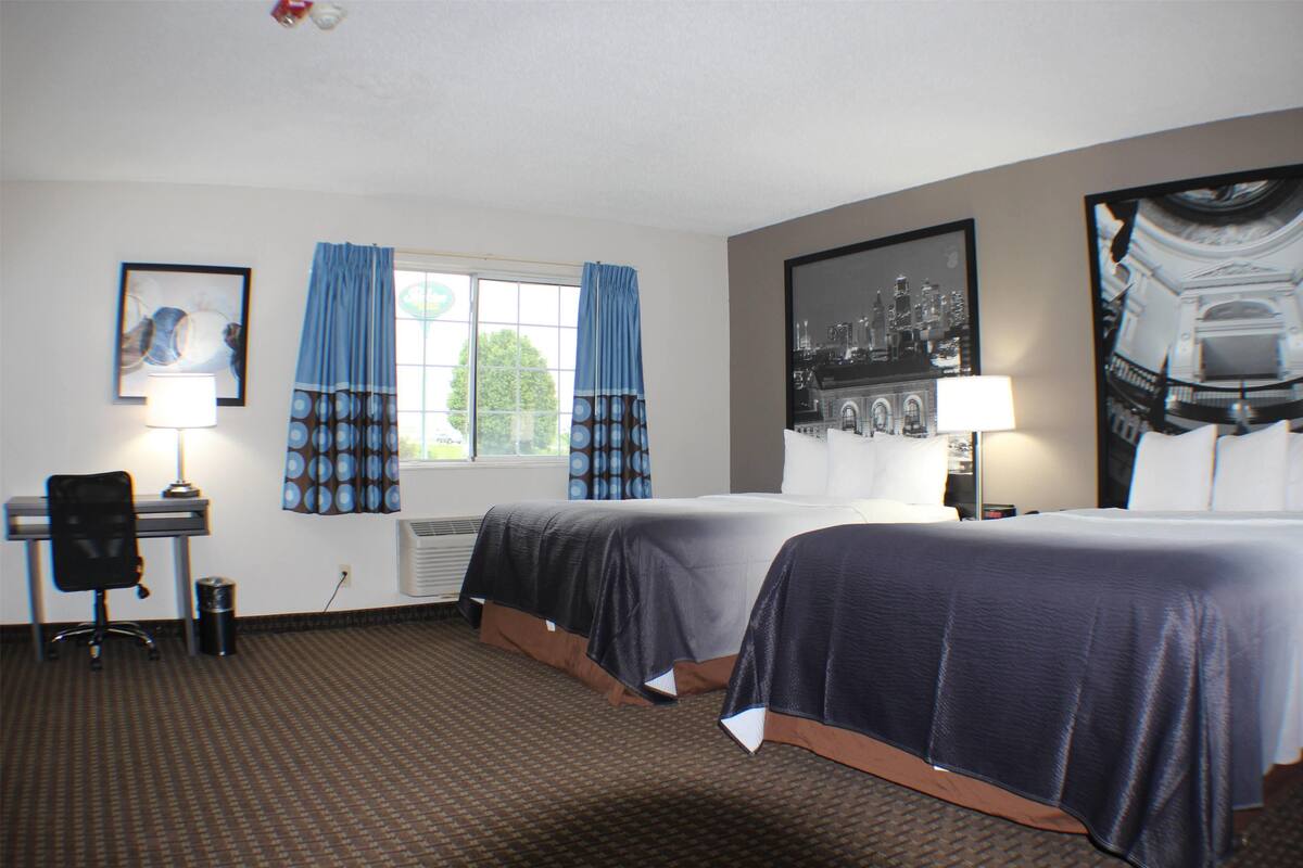 Een hotelkamer met twee bedden, een bureau, een stoel, een prullenbak, een raam met gordijnen en een aan de muur hangend schilderij.