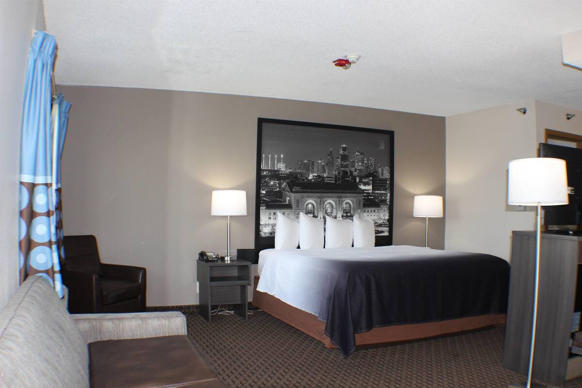 Een hotelkamer met een groot bed, twee nachtkastjes, een bureau, een stoel, een bank en een kunstwerk met een stadsgezicht aan de muur.