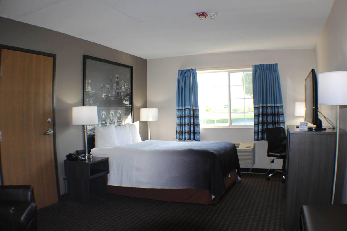 Een hotelkamer met een bed, twee bedlampjes, een bureau met stoel, een raam met gordijnen en een ingelijst kunstwerk aan de muur.