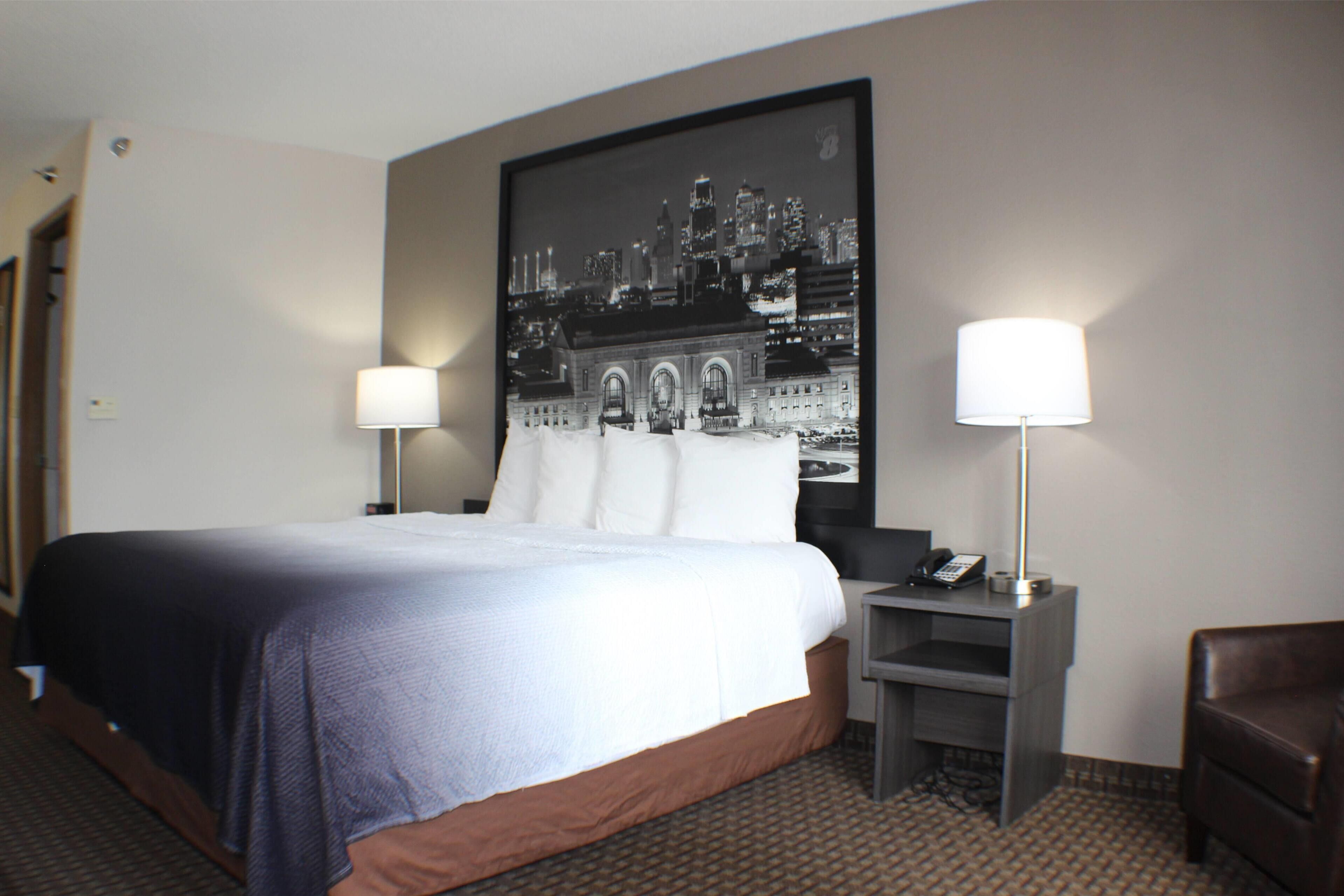 A hotel room with a large bed, two bedside tables with lamps, a chair, and a cityscape artwork on the wall.