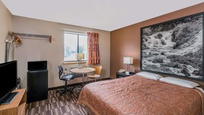 A hotel room with a bed, a desk with a computer, a TV, a refrigerator, a window with curtains, and a large painting on the wall. - Super 8 by Wyndham Madison (Madison)