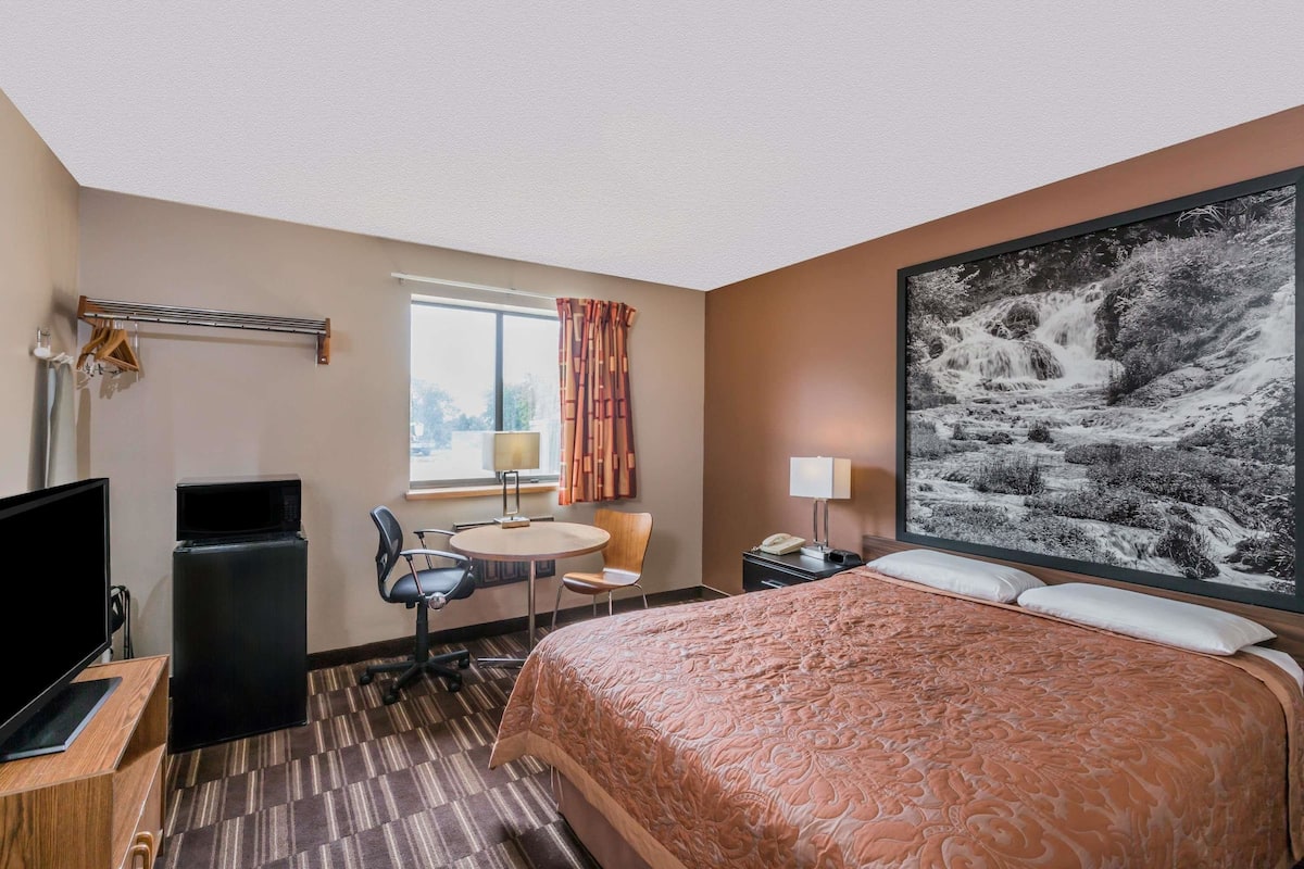 A hotel room with a bed, a desk with a computer, a TV, a refrigerator, a window with curtains, and a large painting on the wall.
