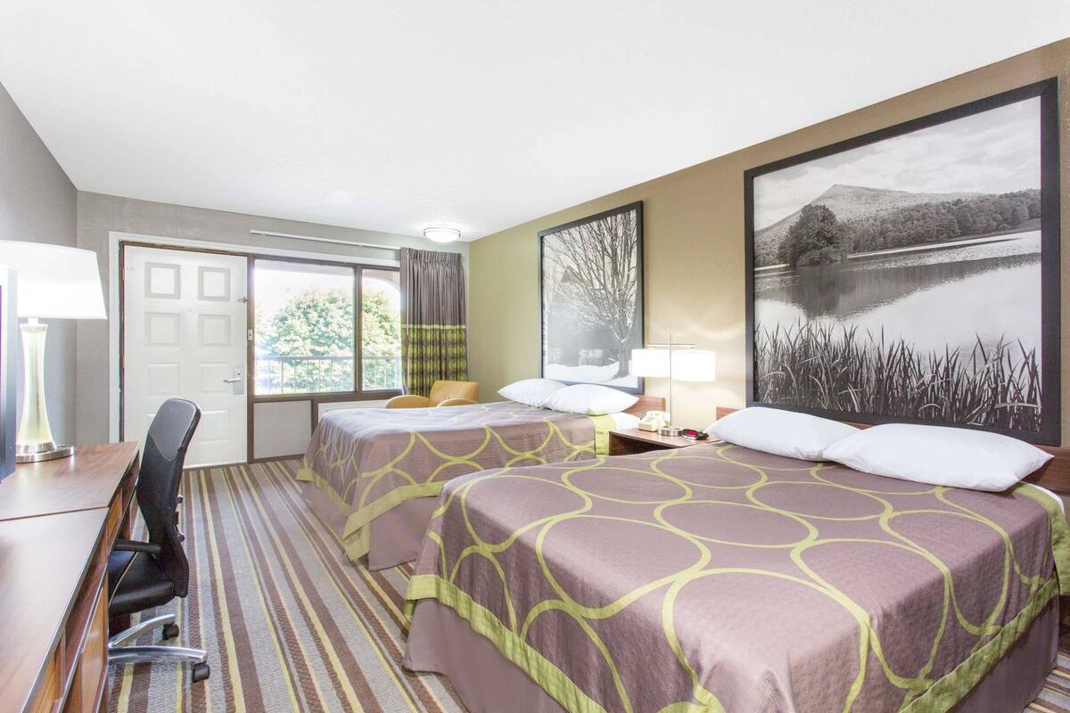 A hotel room with two beds, a desk, a chair, a lamp, and a large window with a view of trees and a lake.