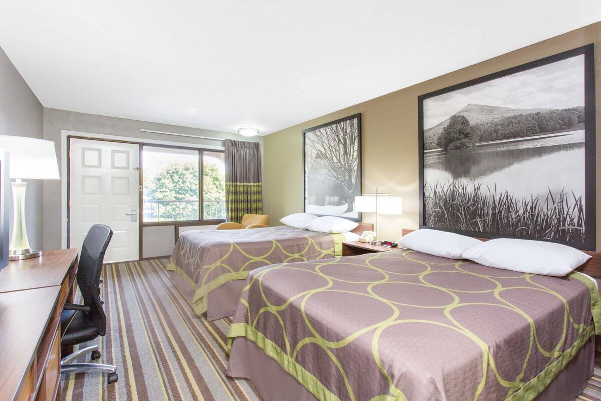 A hotel room with two beds, a desk, a chair, a lamp, and a large window with a view of trees and a lake.