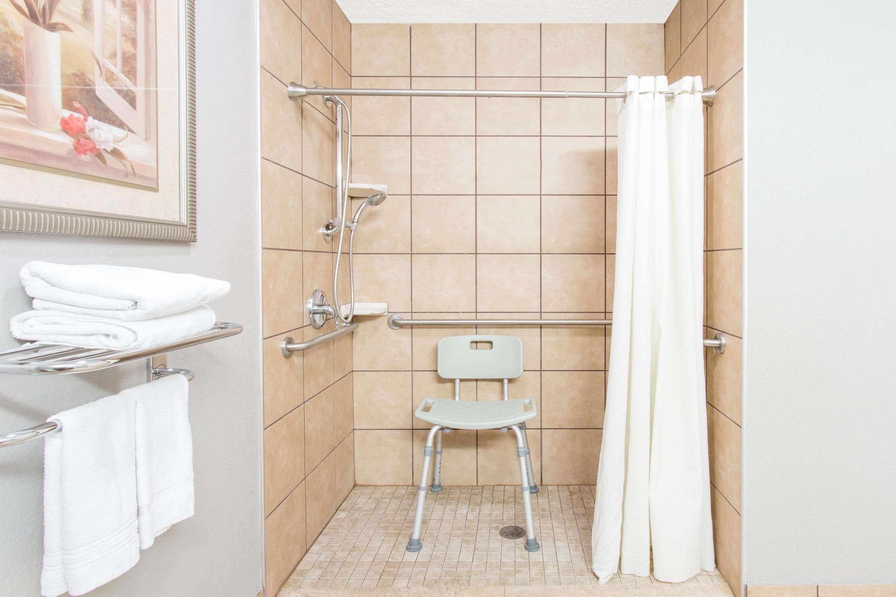 Ein kleines, barrierefreies Duschbad mit Haltegriff, Rollstuhl und weißer Duschvorhang.