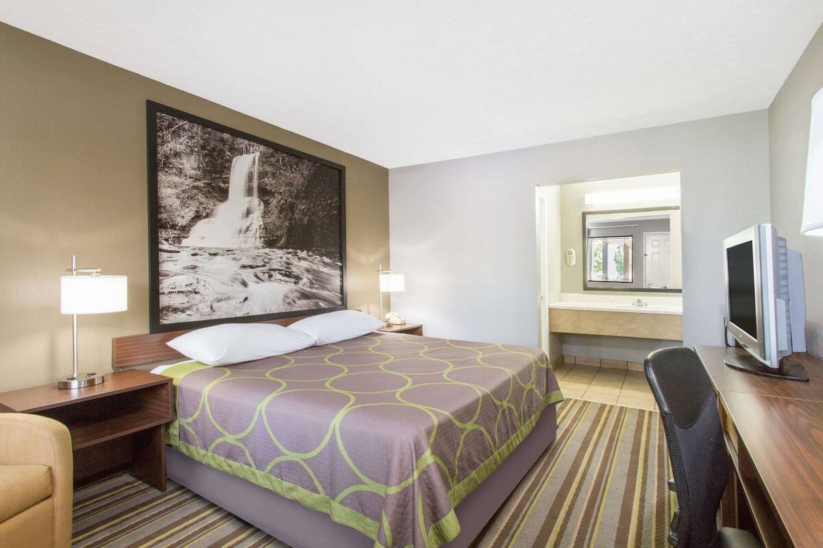 A hotel room with a large bed, a desk with a computer, a TV, a bathroom with a sink and mirror, and a framed picture of a waterfall on the wall.