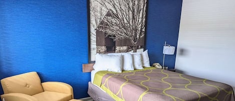 A hotel room with a bed, a chair, a nightstand, and a wall-mounted lamp.