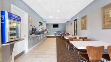 A hotel lobby with a long reception desk, a refrigerator with snacks, a coffee machine, and a TV screen. There are several tables and chairs arranged for dining.
