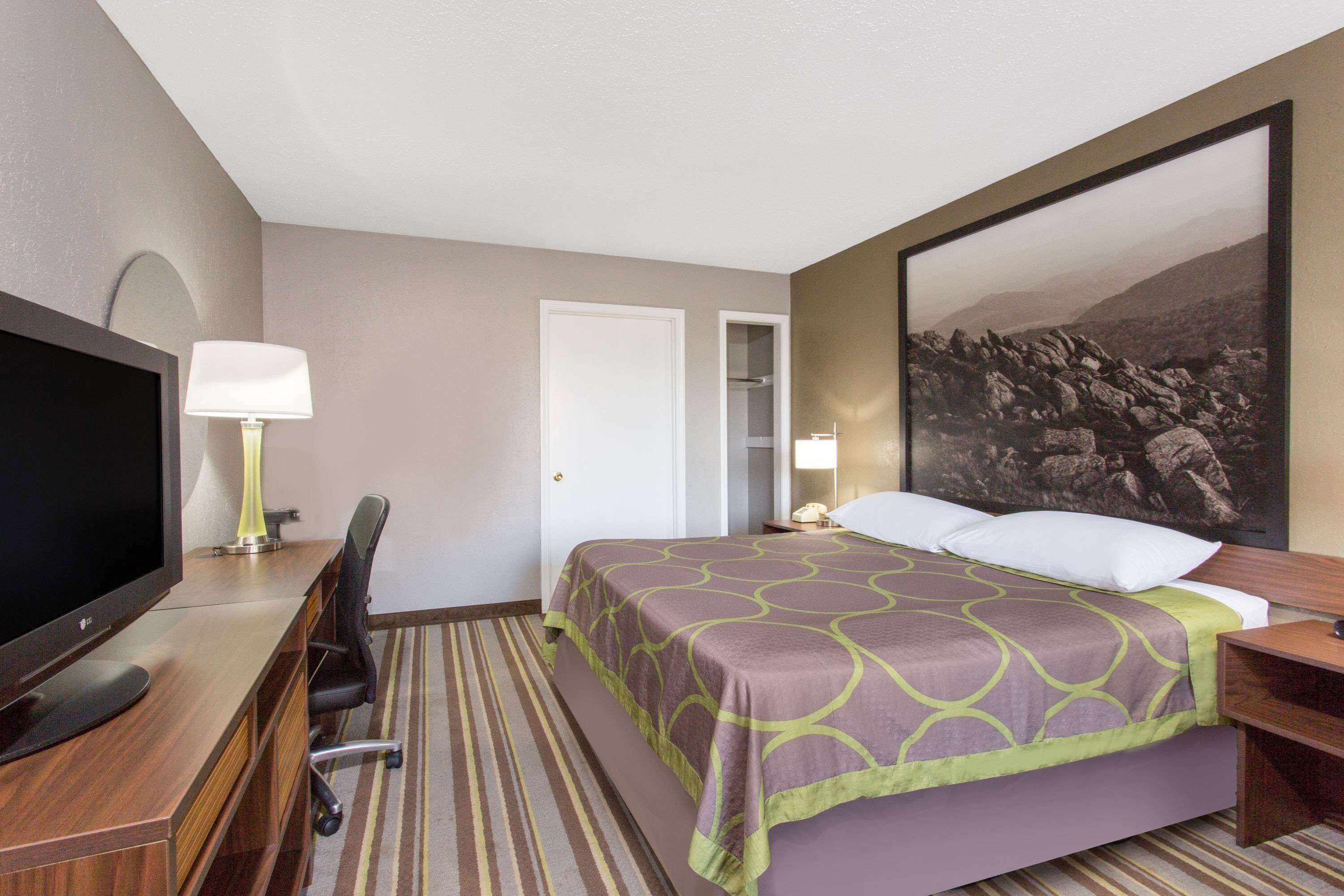 A hotel room with a bed, a desk with a computer monitor, a chair, a lamp, and a large framed picture on the wall.